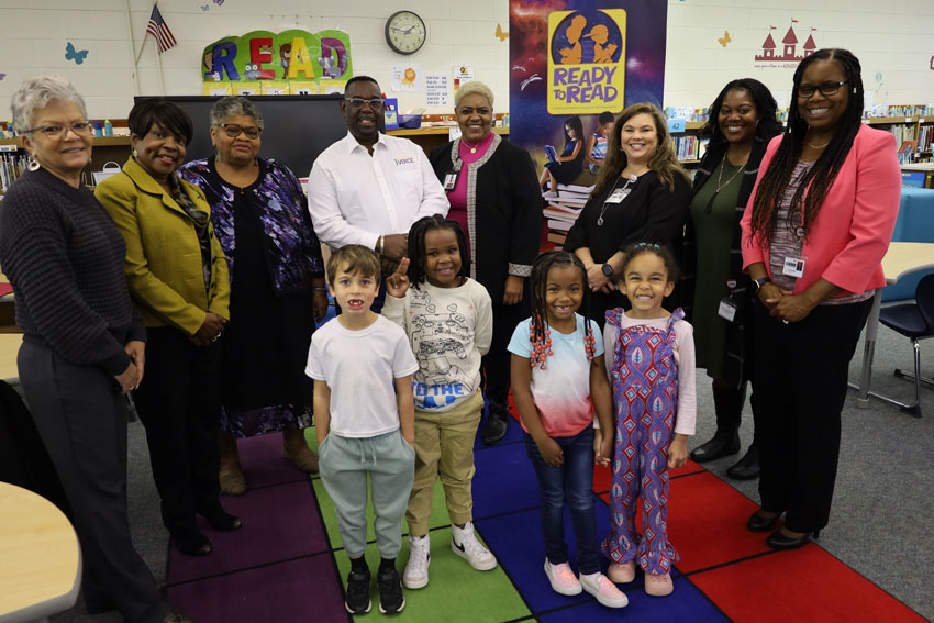 Group of school administrators, community volunteers and students standing in library with "Ready to Read" display in background