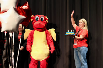 Teacher holding cake next to school mascot--a dragon.