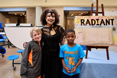 High school drama student in all black costume with two elementary students; "Radiant Reading" display off to side