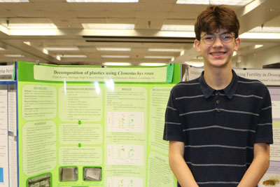 High school student in professional attire standing in front of science fair display with title “Decomposition of Plastics Using Colonostachys rosea”