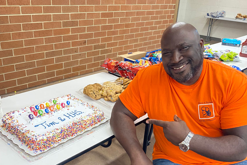 Principal sitting at table pointing to cake that reads "Happy Birthday Tim & HHS"