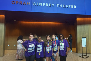 Three students with two teachers in matching t-shirts standing in front of Oprah Winfrey Theater entrance