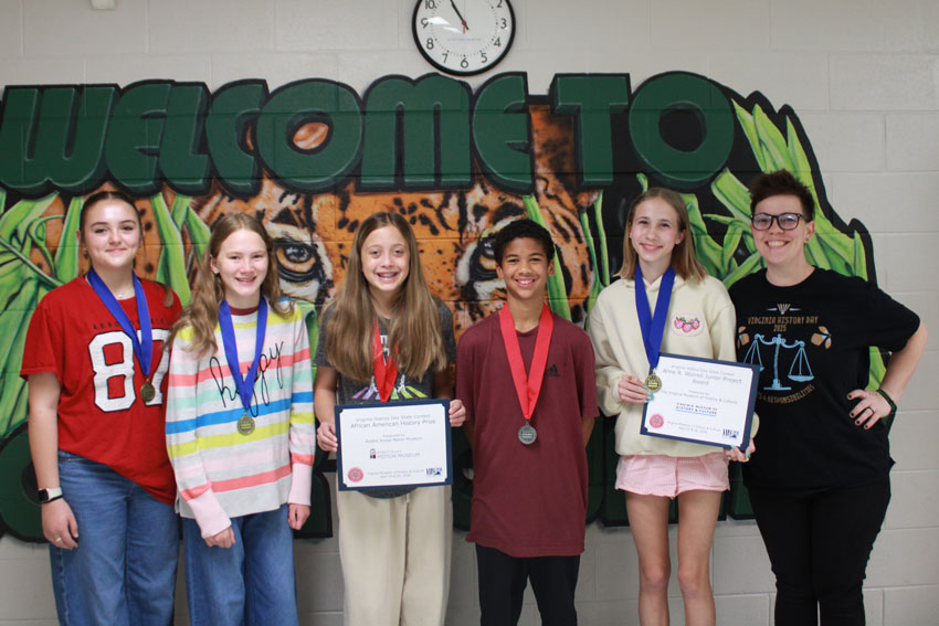 Five middle school students standing with certificates and competition medals standing in front of Linkhorne Middle School mural with their teacher