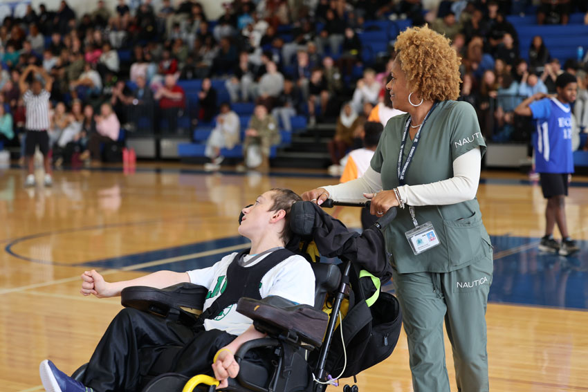 Personal Care Assistant pushing student in wheelchair on basketball court