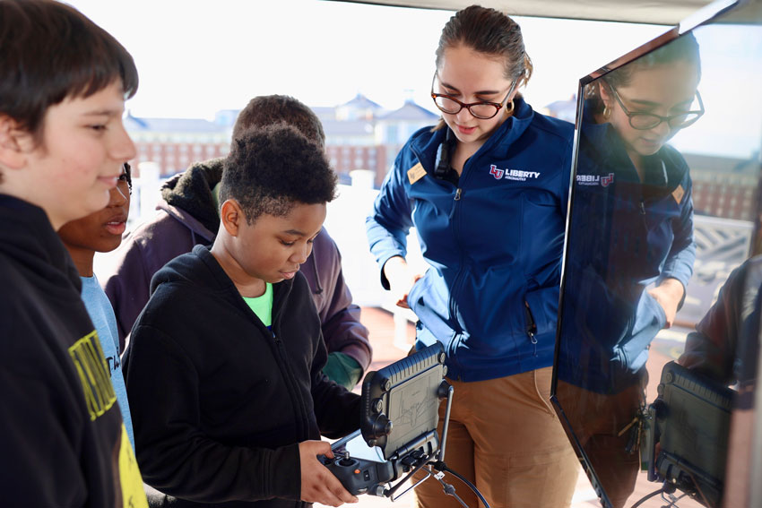 Middle school student holding drone controller with classmates and college student in Liberty University jacket huddled around