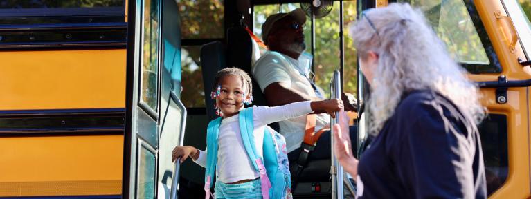 Student with backpack getting off bus with staff member there to greet her