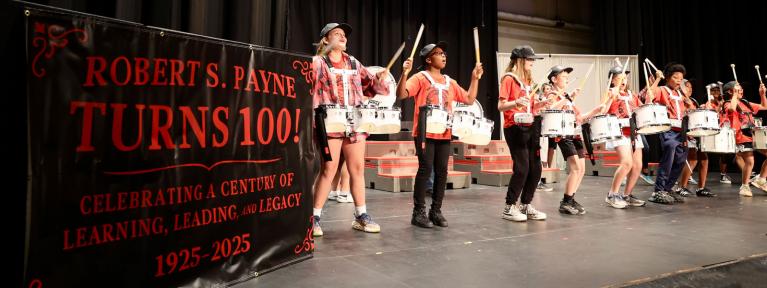 Elementary drum line performing next to banner reading "Robert S. Payne Turns 100!"